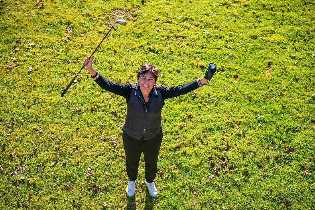 Blick von oben auf eine Golferin auf grünem Gras, die ihren Schläger und Ball in die Luft hält und zur Kamera aufblickt; Schweiz