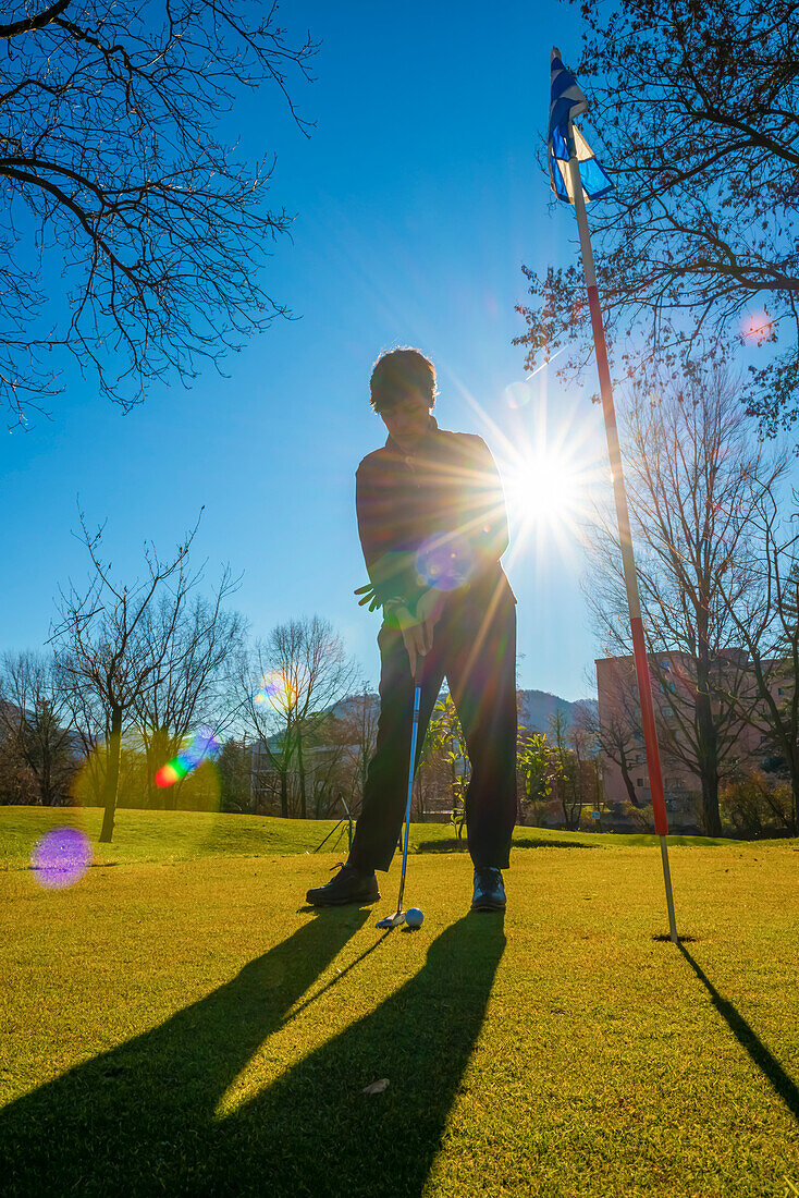 Female golfer using a putter at the hole of a golf course, backlit by sunlight; Switzerland