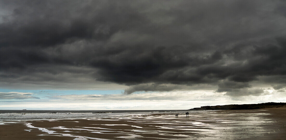 Dark storm clouds over the Atlantic ocean with two people and their dog walking on the wet sand beach in the foreground; South Shields, Tyne and Wear, England