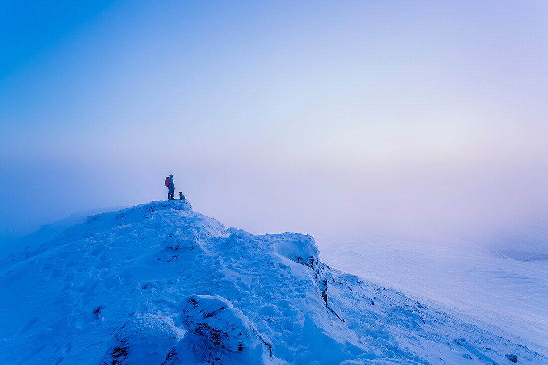 Mann und Hund wandern im Winter auf einem schneebedeckten Berg im Nebel, Galty Mountains; County Tipperary, Irland