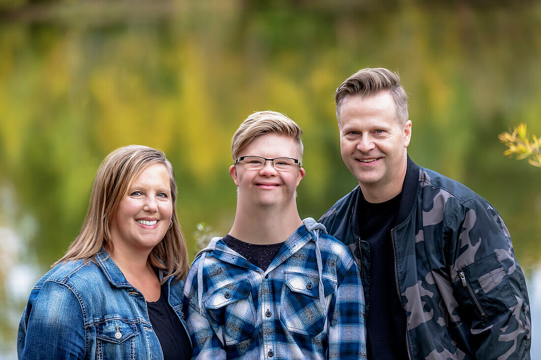 A young man with Down Syndrome posing for a family portrait with his father and mother while enjoying each other's company in a city park on a warm fall evening: Edmonton, Alberta, Canada
