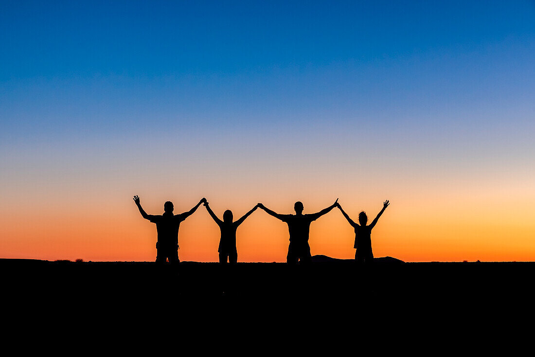 Silhouette von vier Menschen, die sich bei Sonnenuntergang mit erhobenen Armen an den Händen halten in Aluvlei, Namib-Naukluft-Nationalpark; Namibia