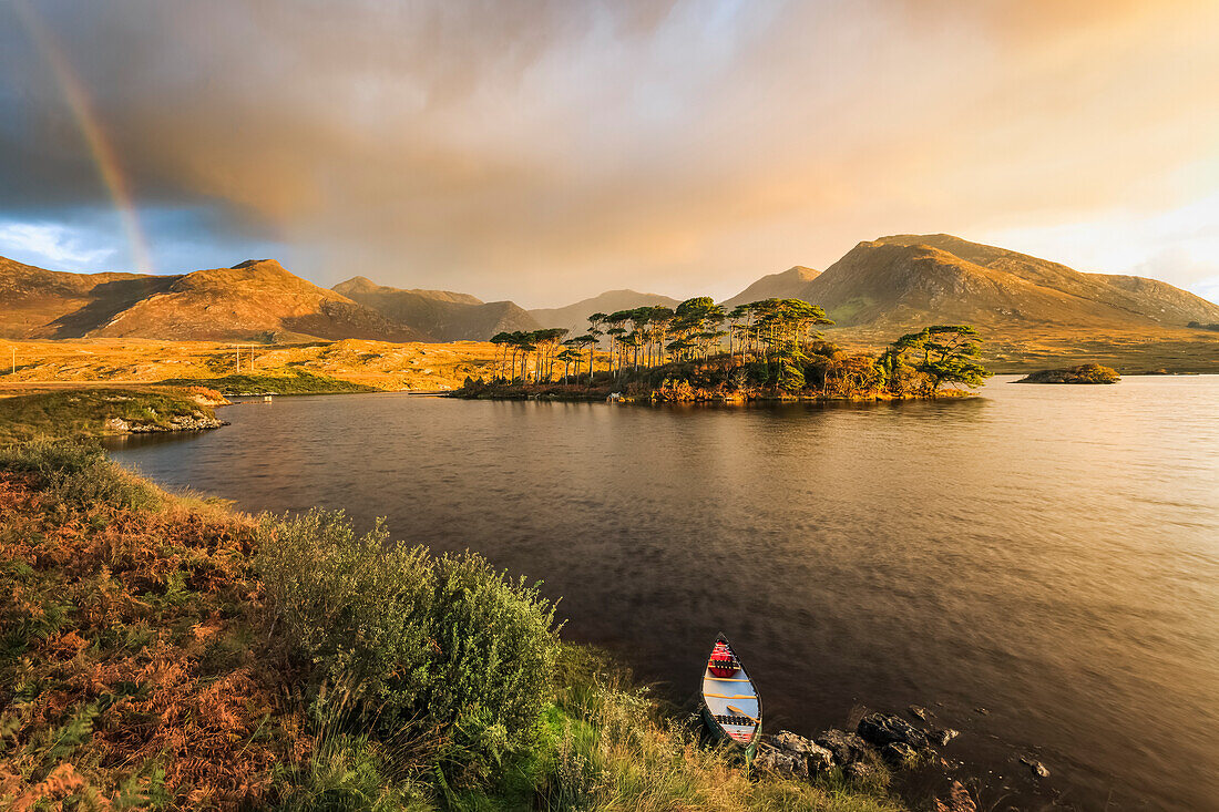Canoe on the banks of a lake in Connemara with an island with trees at sunrise with storm clouds and a rainbow in the distance; Connemara, County Galway, Ireland