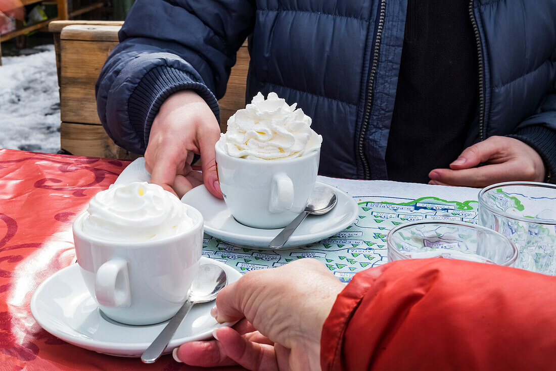 Ein Paar in Skijacken sitzt an einem Tisch und bekommt Tassen mit heißer Schokolade und Schlagsahne serviert; Courmayeur, Aostatal, Italien