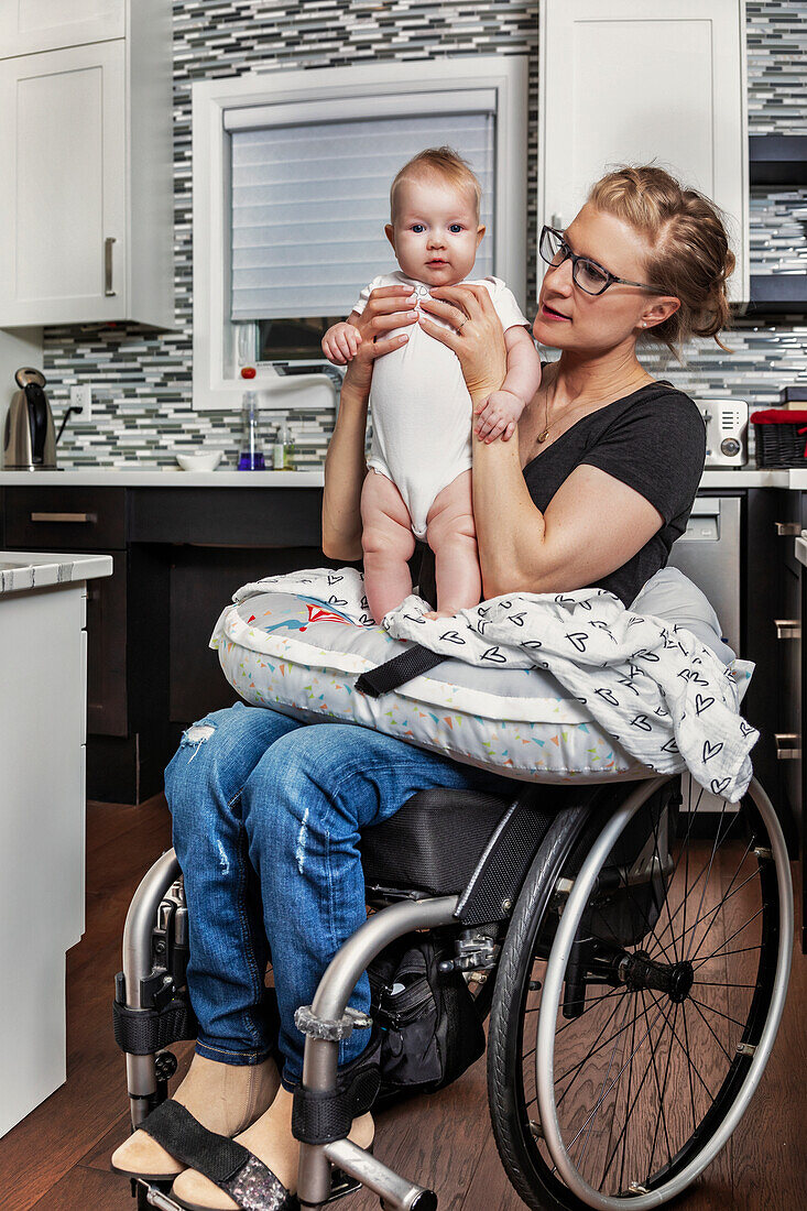 Eine querschnittsgelähmte Mutter hält ihr Baby in der Küche auf dem Schoß, während sie im Rollstuhl sitzt: Edmonton, Alberta, Kanada
