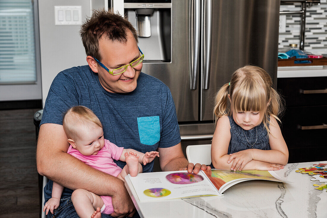 A father sitting down with his daughter in the kitchen to read a book and build a puzzle while holding his baby in his lap: Edmonton, Alberta, Canada