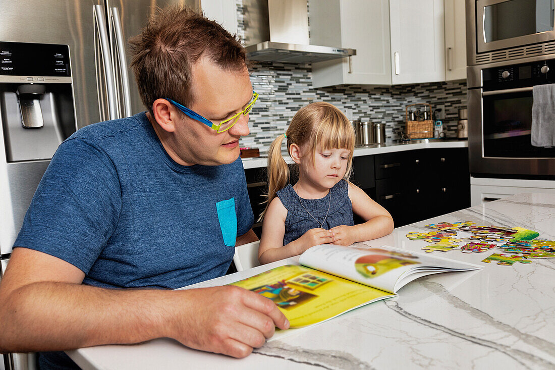 A father sitting down with his young daughter in the kitchen to read a book: Edmonton, Alberta, Canada