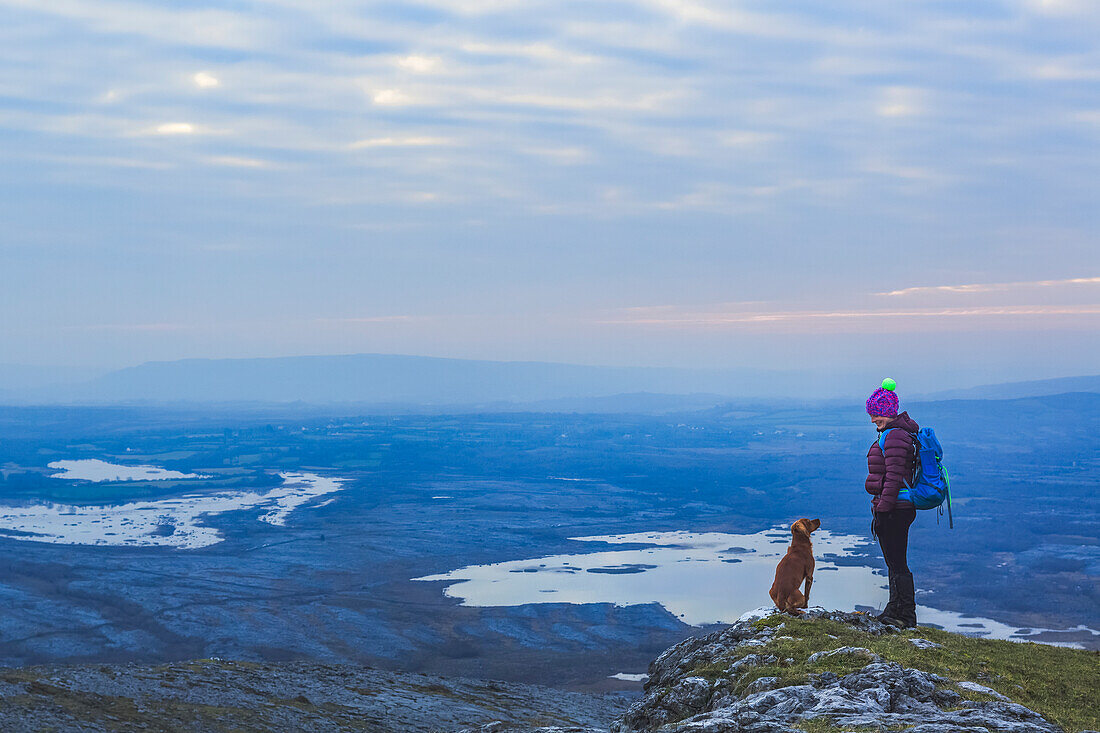 Einsame Wanderin mit Strickmütze und Hund, die sich an einem bewölkten Abend im Winter auf einer Klippe mit Blick auf Seen in der Ferne ansehen, Burren National Park; County Clare, Irland