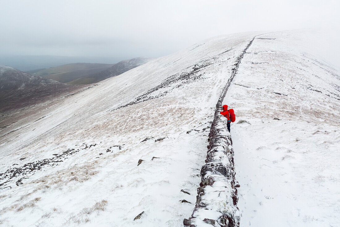 Wanderin blickt über eine alte Steinmauer auf dem Gipfel der Galty Mountains im Winter bei bewölktem, nebligem Wetter; County Tipperary, Irland