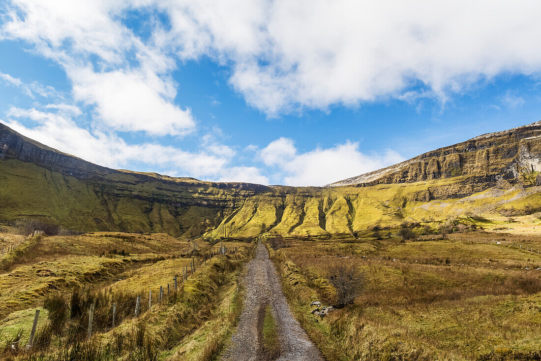 Old Irish gravel road leading to steep cliff and mountains in the sunlight on a sunny day with clouds in the sky; Eagles Rock, County Leitrim, Ireland