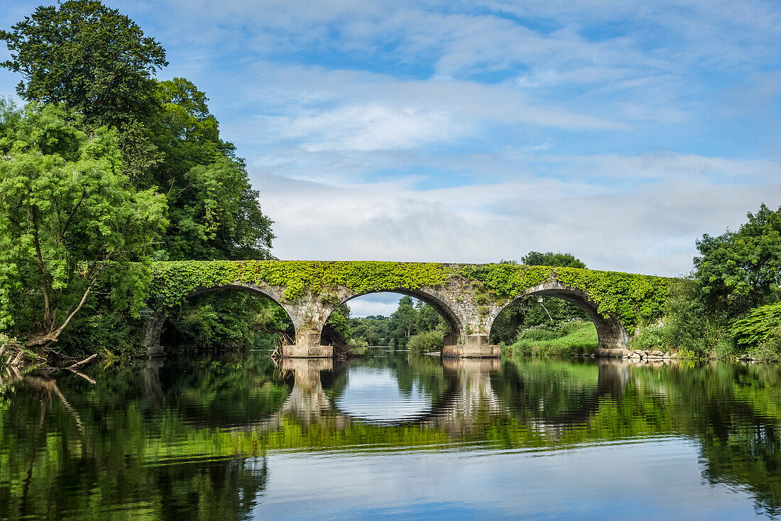Old stone bridge over the Blackwater river in Kilavullen reflected in the water on a sunny summer day; Killavullen, County Cork, Ireland