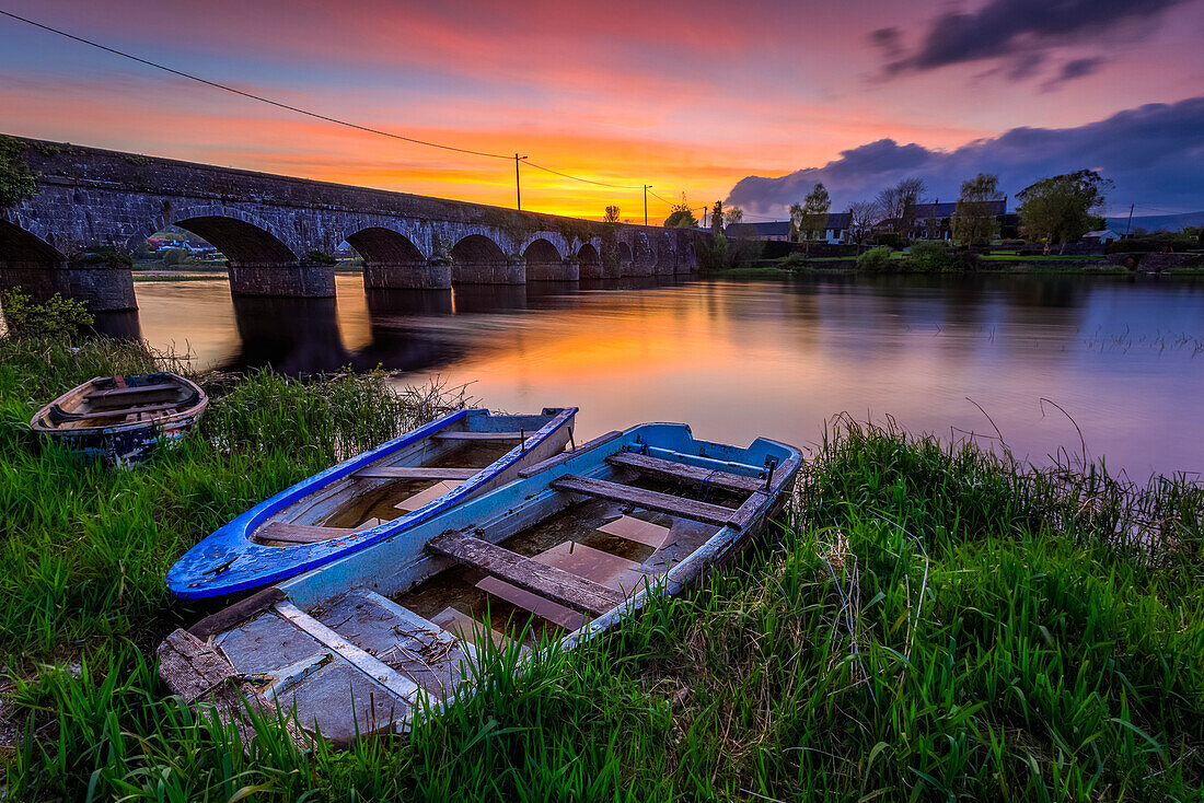 Zwei kleine Holzboote am Ufer des Flusses Shannon vor einer Steinbrücke bei Sonnenuntergang im Sommer; O'Brien's Bridge, County Clare, Irland