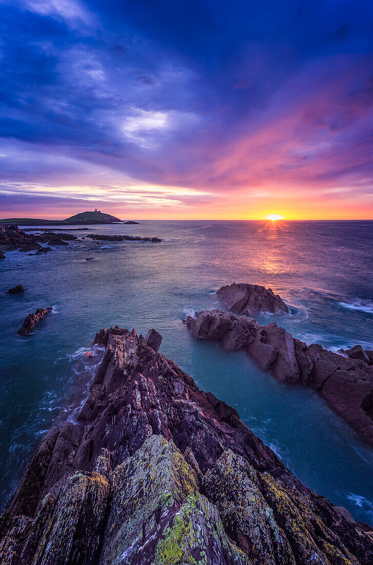 Sunrise along the Irish coast with rock formations in the foreground and Ballycotton lighthouse on the horizon; Ballycotton, County Cork, Ireland