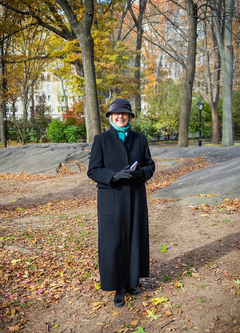 Ältere Frau steht für ein Porträt im Central Park im Herbst, hält ein Smartphone und trägt einen langen Mantel; New York City, New York, Vereinigte Staaten von Amerika