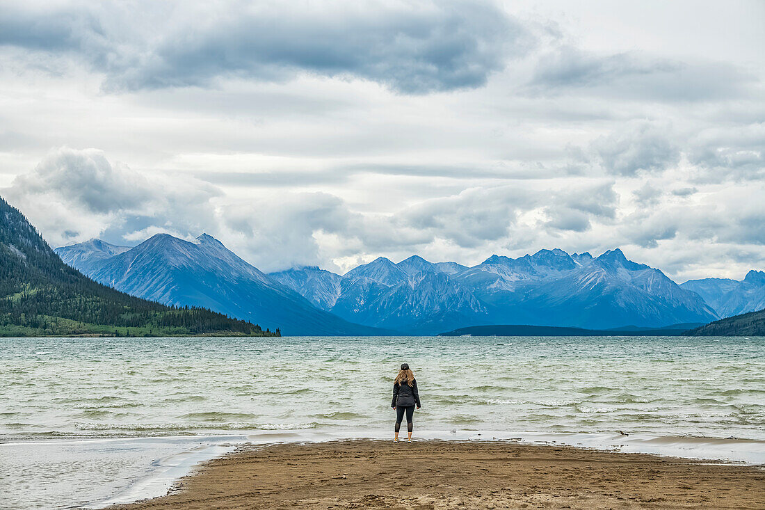 Frau steht an einem Sandstrand und blickt auf den Carcross Lake und die gewaltigen Yukon Ranges; Carcross, Yukon, Kanada