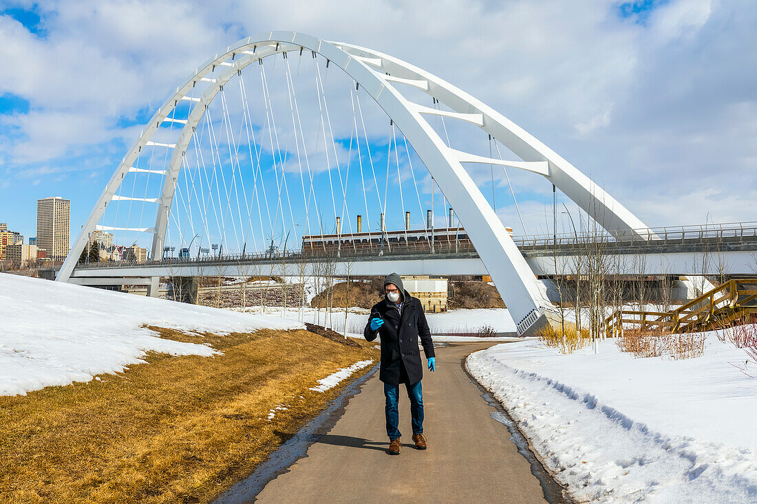 Ein Mann mit Maske und Handschuhen geht auf einem Weg im Freien und benutzt sein Smartphone während der Covid-19-Weltpandemie; Edmonton, Alberta, Kanada