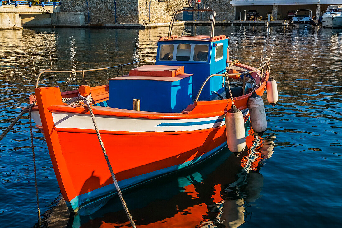 Small traditional orange, white and blue Greek fishing boat with boat fenders docked in Mykonos new port marina at sunrise; Mykonos Town, Mykonos Island, Greece