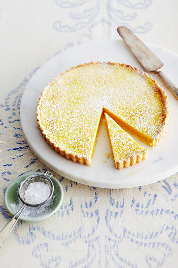 Lemon Tart on Marble Stand with Pie Server and Bowl of Icing Sugar on Table in Studio