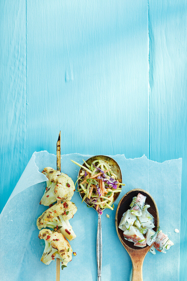 Draufsicht auf gegrillten Blumenkohl am Spieß und mit Salat gefüllte Löffel auf Wachspapier auf blauem Holztisch im Studio
