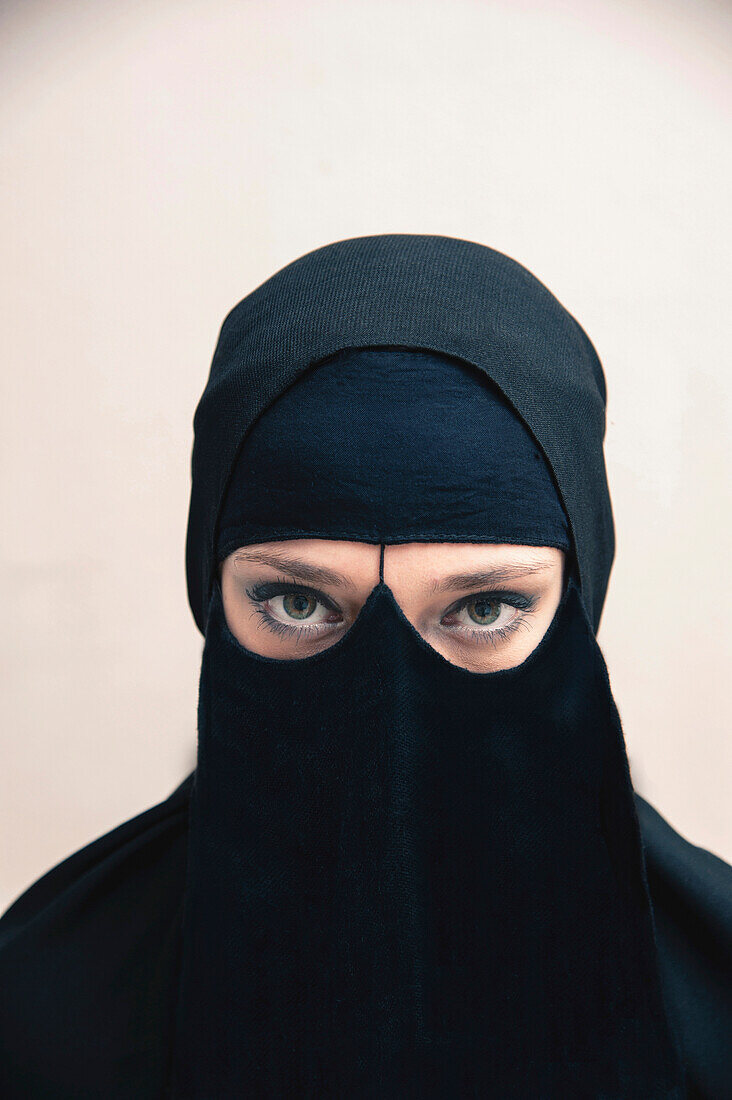 Close-up portrait of young woman wearing black, muslim hijab and muslim dress, eyes looking at camera showing eye makeup, studio shot on white background