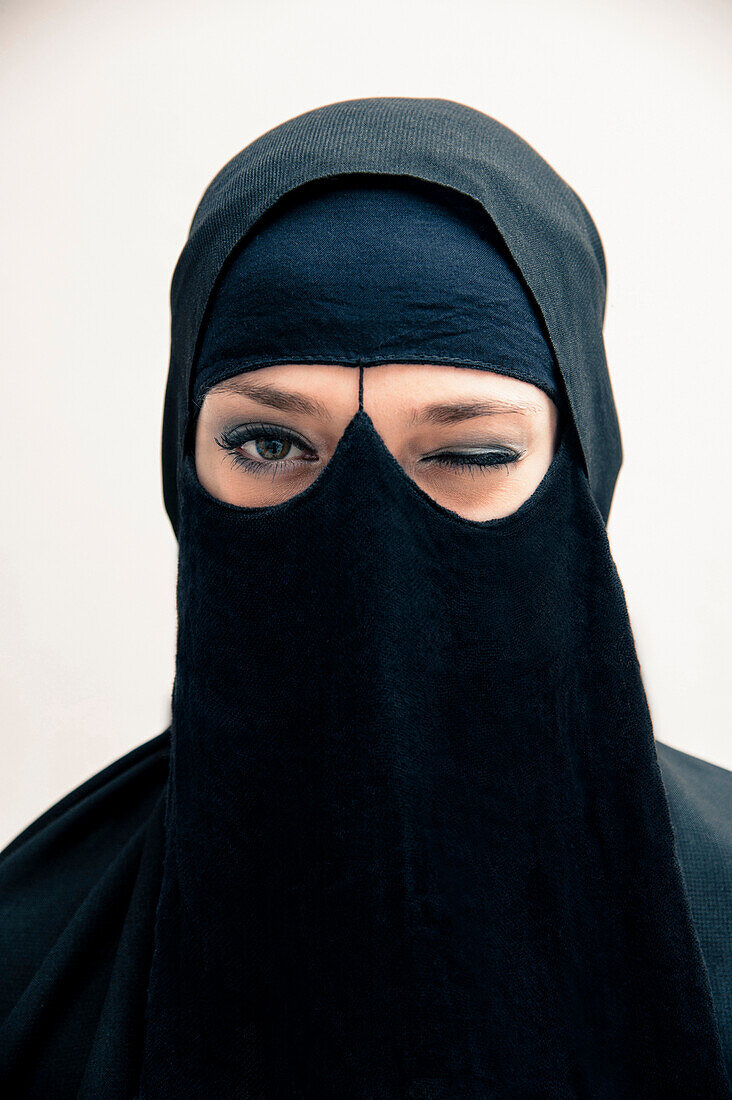 Close-up portrait of young woman wearing black, muslim hijab and muslim dress, winking and looking at camera, eyes showing eye makeup, studio shot on white background