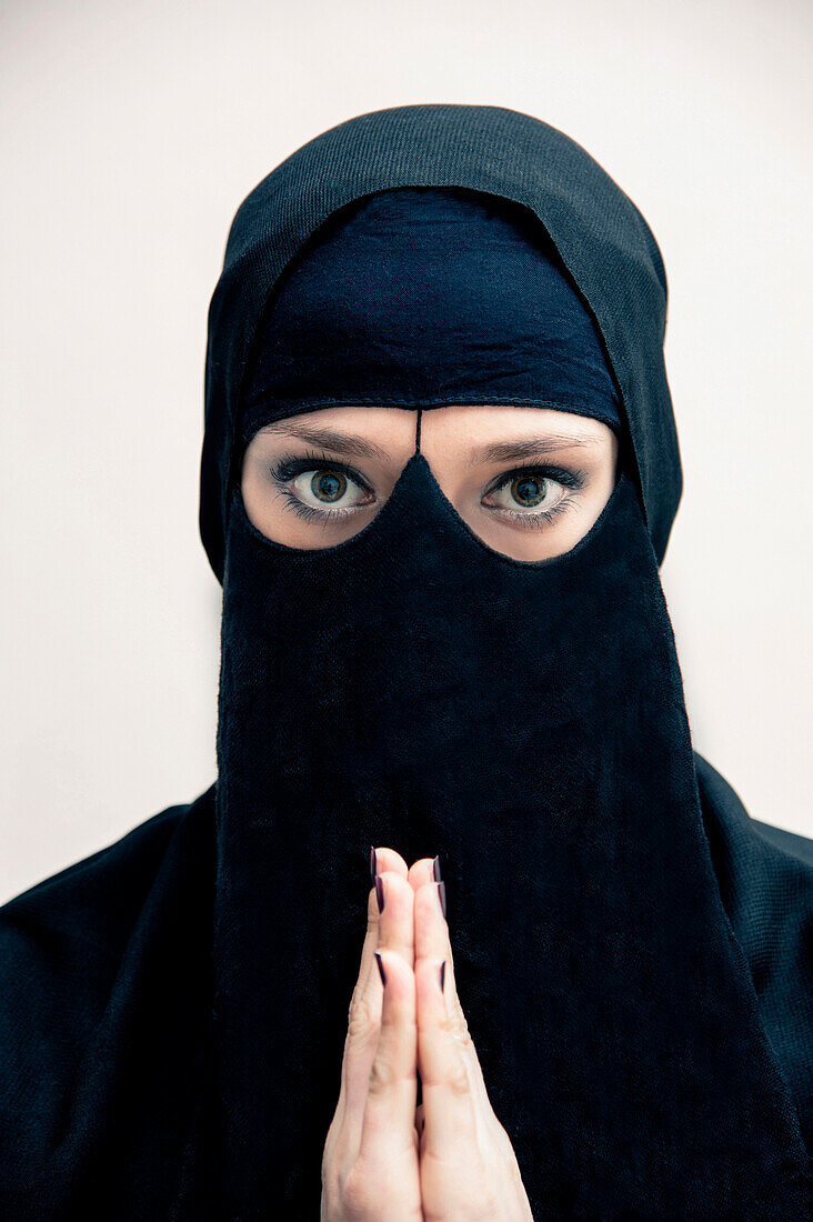Close-up-Porträt einer jungen Frau in schwarzem muslimischem Hijab und muslimischem Kleid, die mit den Händen betet und in die Kamera schaut, mit geschminkten Augen, Studioaufnahme auf weißem Hintergrund