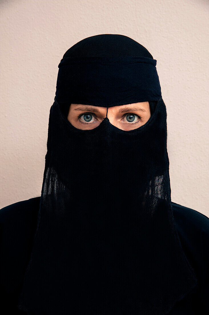 Nahaufnahme einer Frau, die einen schwarzen muslimischen Hidschab und ein muslimisches Kleid trägt und in die Kamera schaut, Studioaufnahme auf weißem Hintergrund