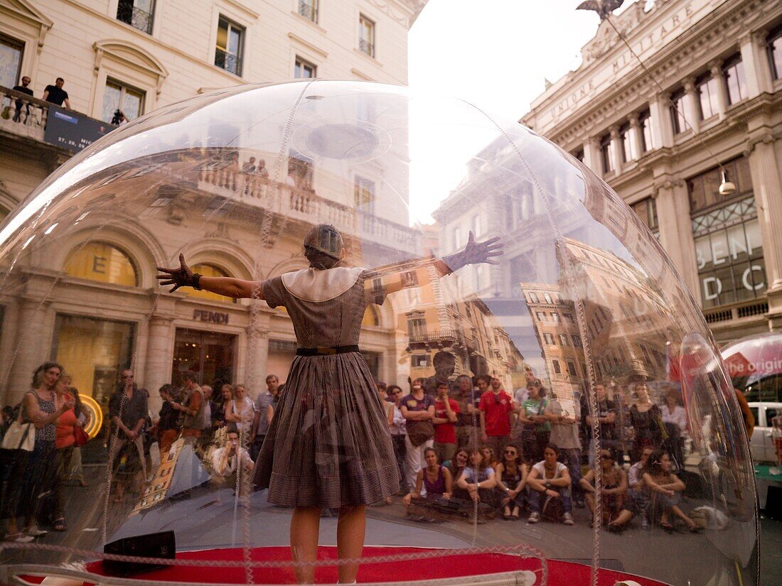 Frau, die in einer durchsichtigen Glaskugel steht, und eine Menschenmenge, die ihre Darbietung beobachtet; Rom, Italien