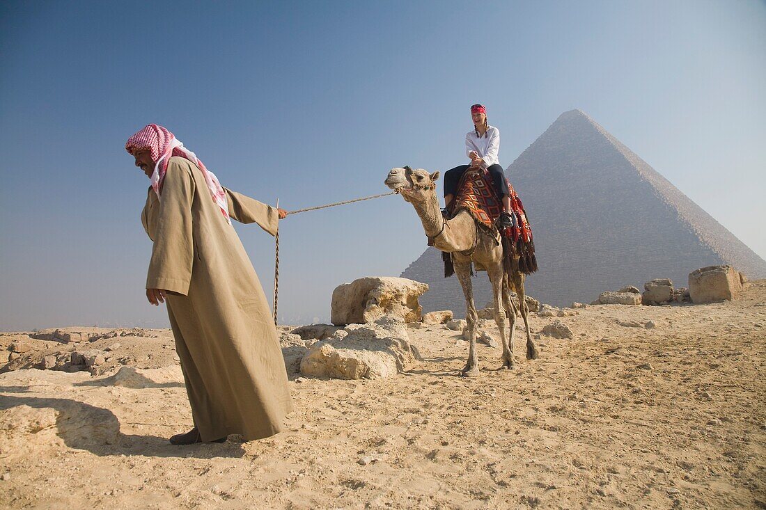 Young Woman Tourist Riding A Camel Lead By A Guide At The Pyramids Of Giza, Egypt