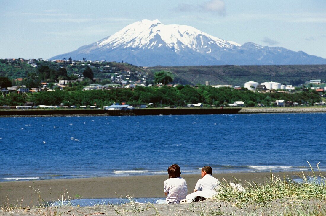 Couple Sitting On Shore By Snow-Covered Mountain And Lake