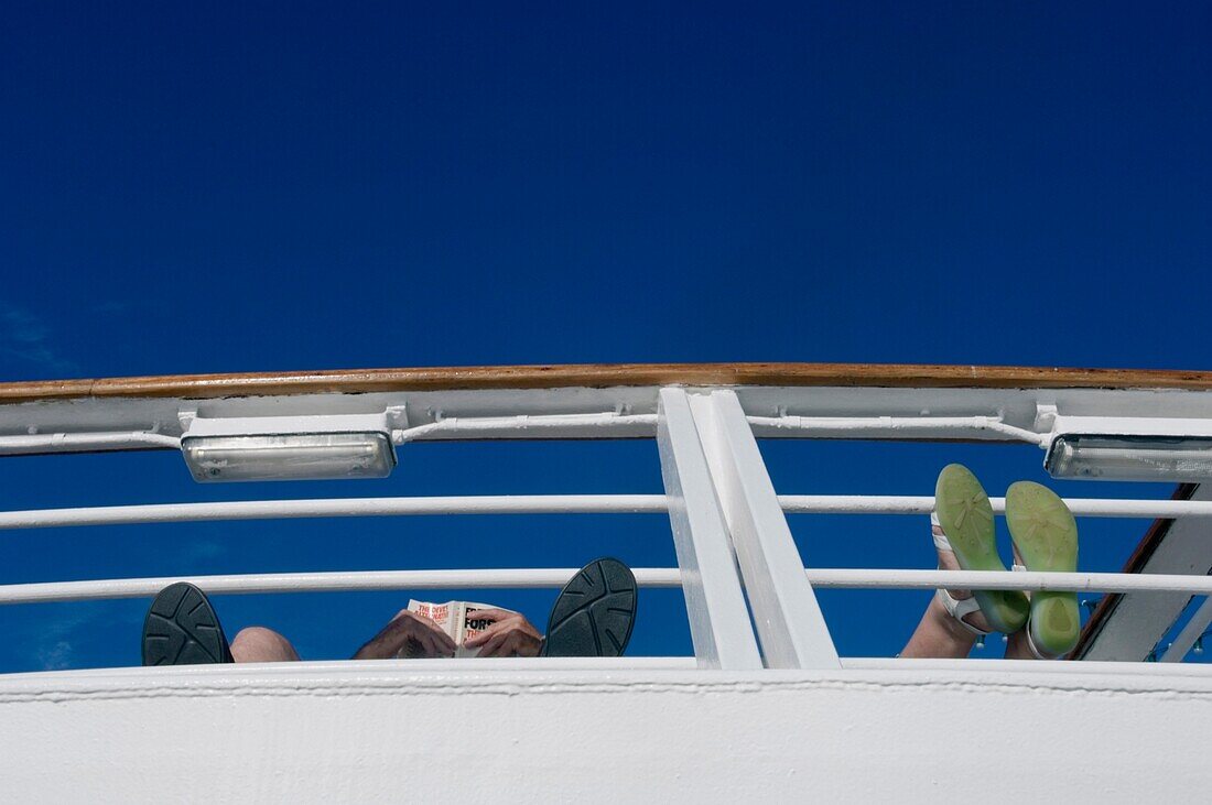 Passagiere an Bord eines Kreuzfahrtschiffes lesen mit den Füßen auf der Reling