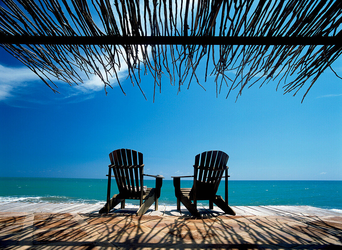 Zwei Stühle auf Deck am Meer im Schatten eines Grasdachs