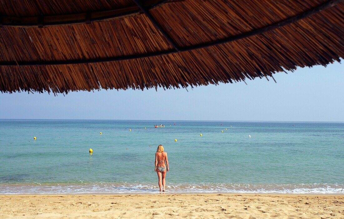 Blick durch einen mit Gras bedeckten Sonnenschirm auf eine Frau am Strand