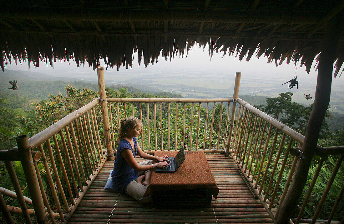 Eine Frau, die auf dem Boden sitzt und einen drahtlosen Laptop-Computer benutzt, um E-Mails abzurufen, auf dem Balkon einer abgelegenen Bambushütte mit Blick auf Bangladesch