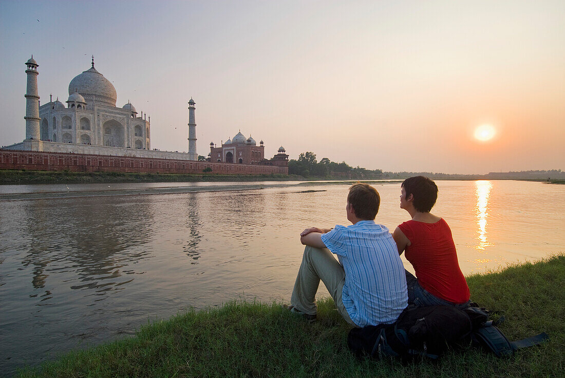 Paar, das auf einem Rucksack sitzt und in der Abenddämmerung den Yamuna-Fluss überblickt und das Taj Mahal bewundert