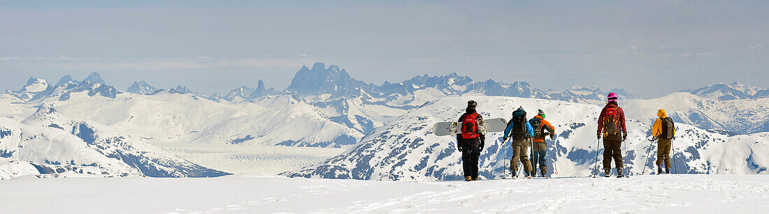 Ein Snowboarder und Skifahrer wandern beim Heliskiing in der Nähe von Juneau, Alaska, entlang des Westgrats des Mt. Hawthorne mit Blick auf das Juneau-Eisfeld und die Teufelskralle