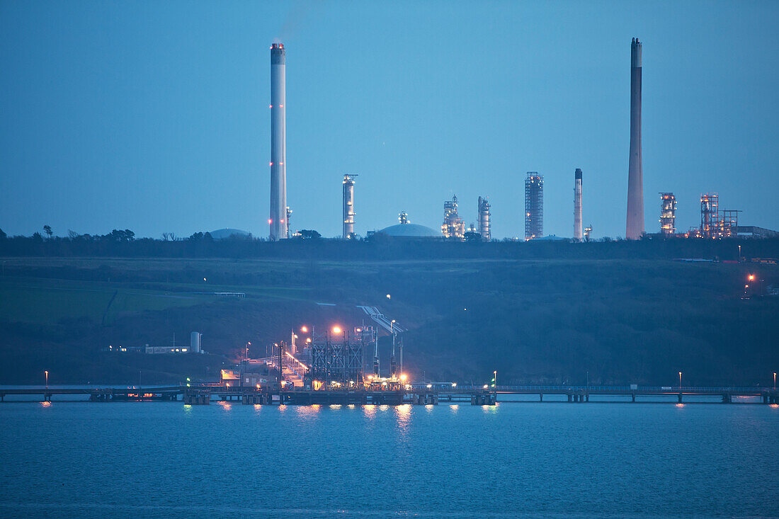 Lichter beleuchten eine Struktur auf einem Pier über dem Wasser mit Schornsteinen im Hintergrund