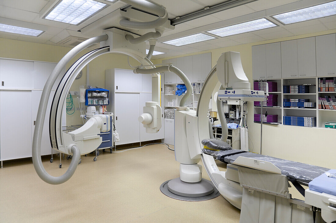 A modern hospital room, a large portable mobile scanning machine with curved shaped arms, a mobile scanner and hospital gurney or bed.