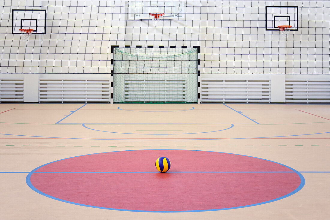 Eine Schulsporthalle mit einem markierten Hallenfußballfeld, Toren und Basketballkörben, einem blau-gelb gestreiften Ball in einem rosa Kreis. 
