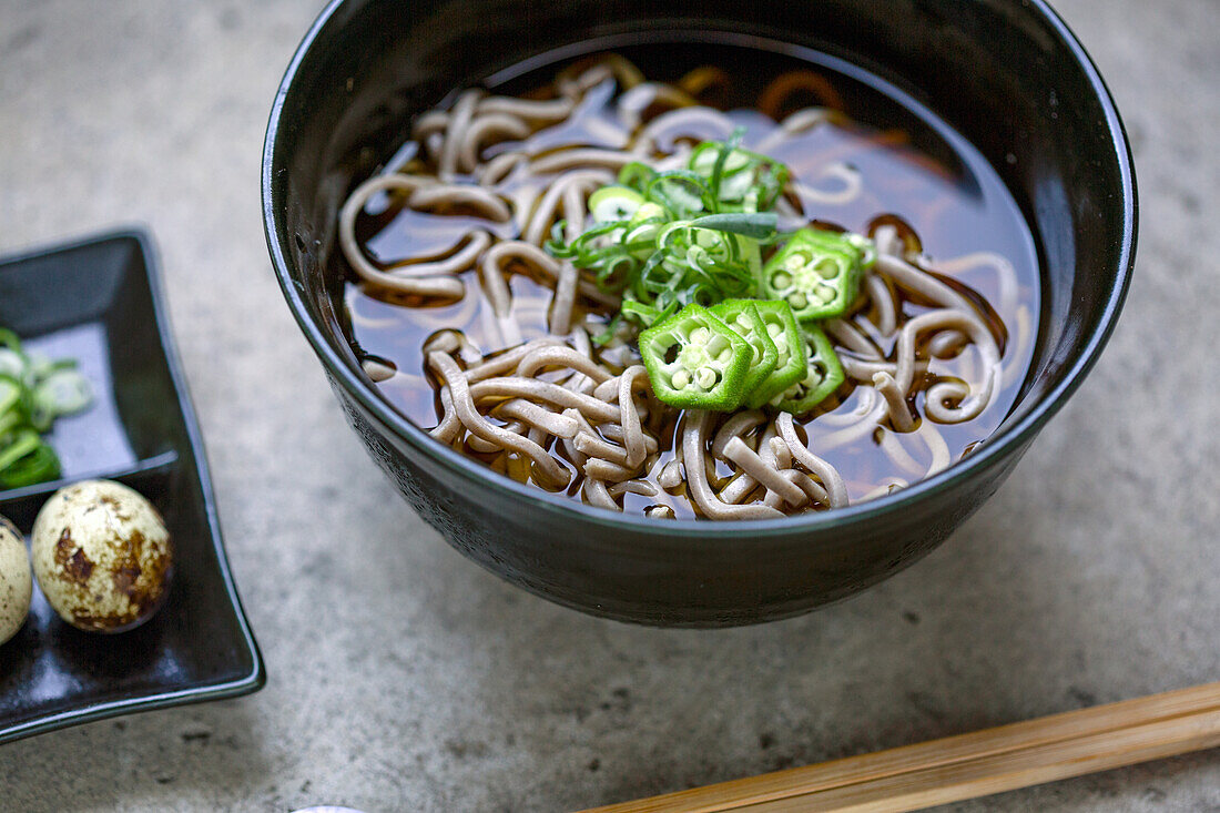 A black china bowl of noodles and broth, sliced green chilli and a dish with quail's eggs and sliced vegetables.