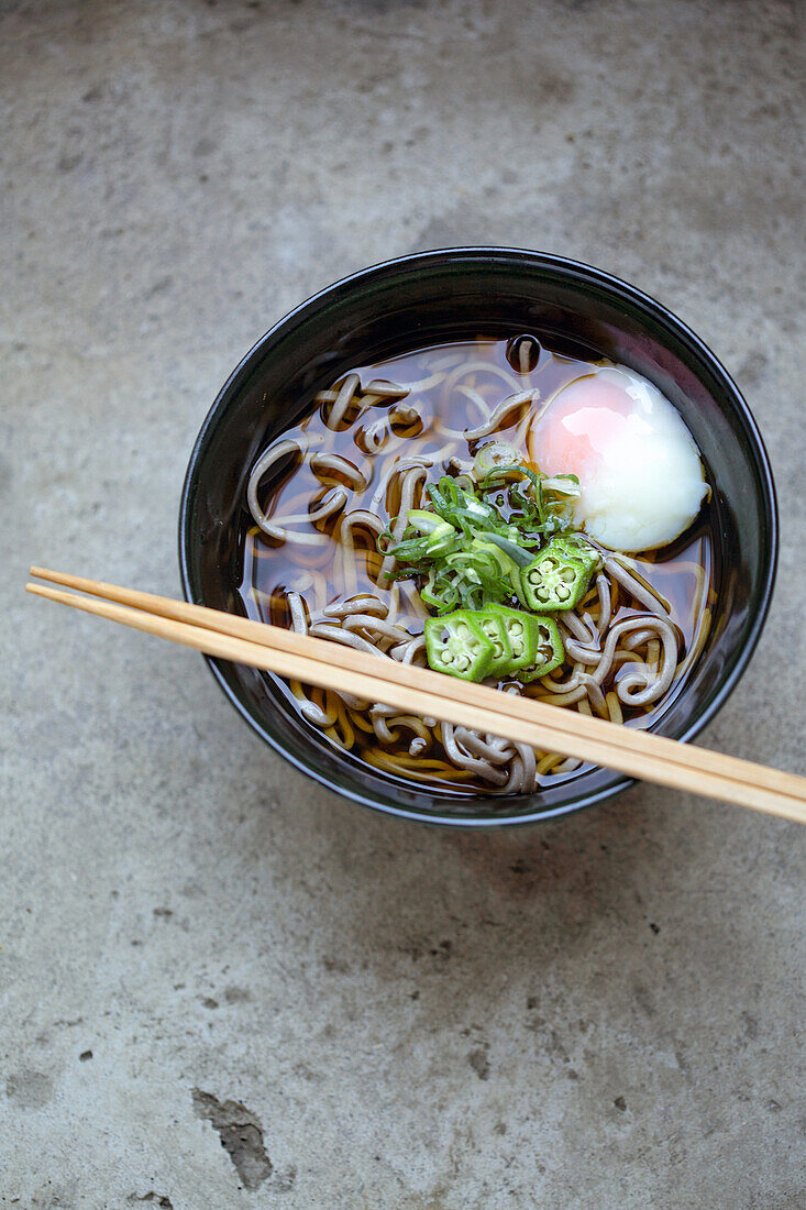 A black china bowl of noodles and broth with sliced chillis and an egg.