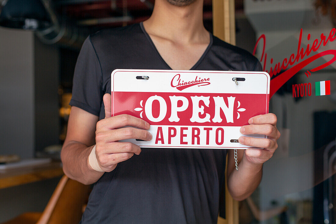 Man hanging up an Open sign on a restaurant door. Dual language, Italian and English. 
