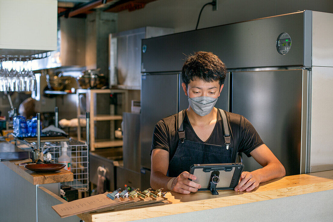 Ein Mann mit einer Gesichtsmaske an der Theke einer Restaurantküche, der ein digitales Tablet benutzt, der Eigentümer oder Manager. 