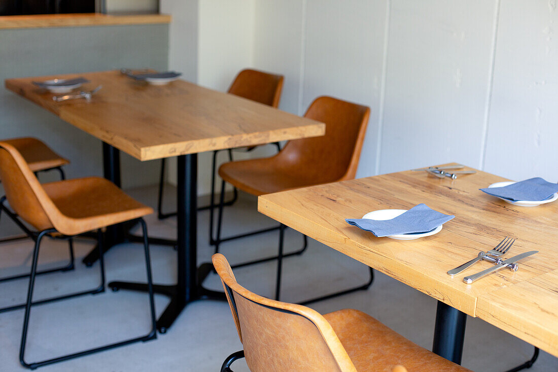 Tables and chairs in a restaurant, plain white walls and grey flooring, place settings. 