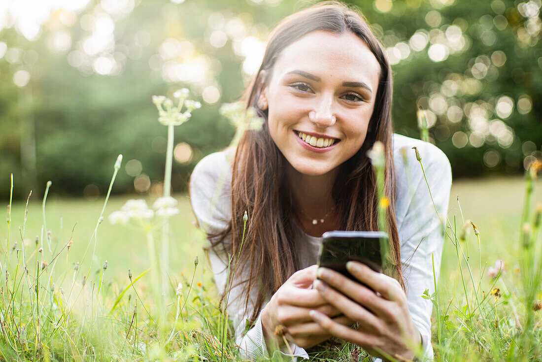 Porträt einer jungen Frau, die ihr Smartphone benutzt, während sie im Park liegt