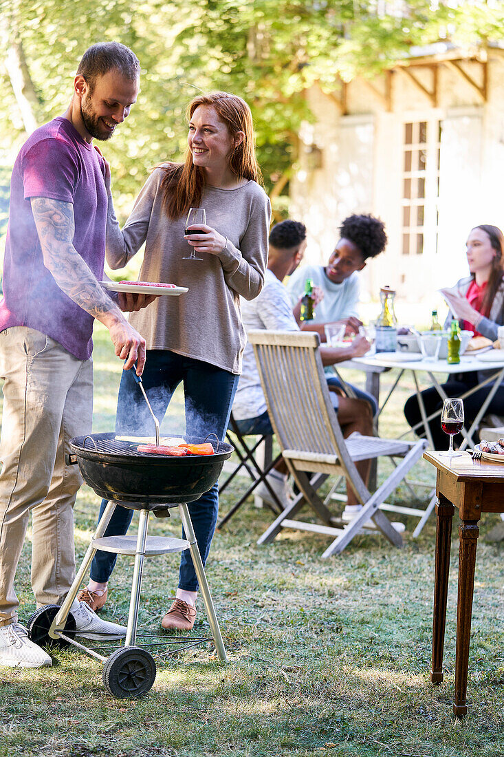 Junger Mann bereitet Essen auf dem Grill vor, während seine Freunde im Hintergrund am Tisch sitzen