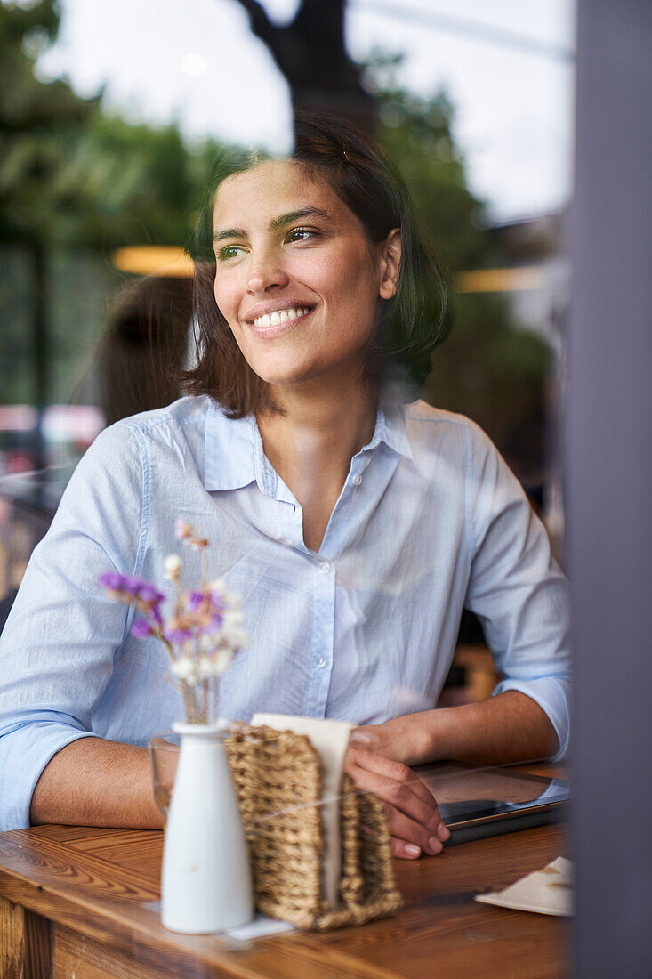 Porträtaufnahme einer lächelnden lateinamerikanischen Frau, die in einem Café sitzt, durch ein Fenster aufgenommen