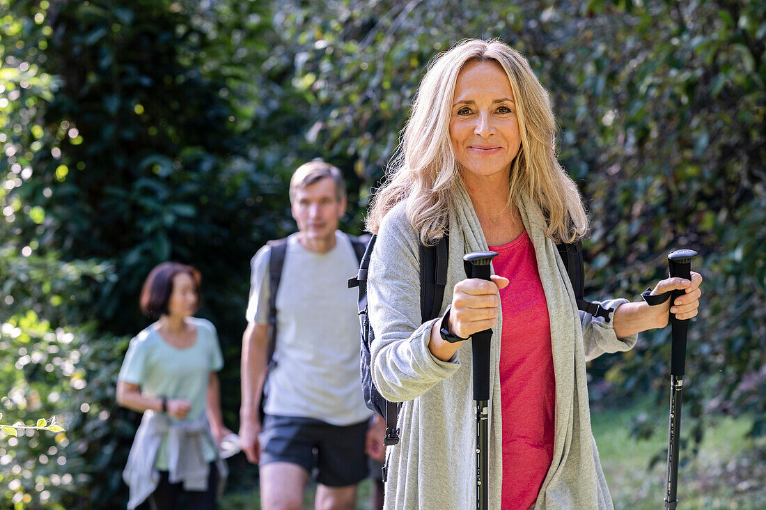 Blonde senior lady carrying backback and hiking poles hiking in the woods with group of friends