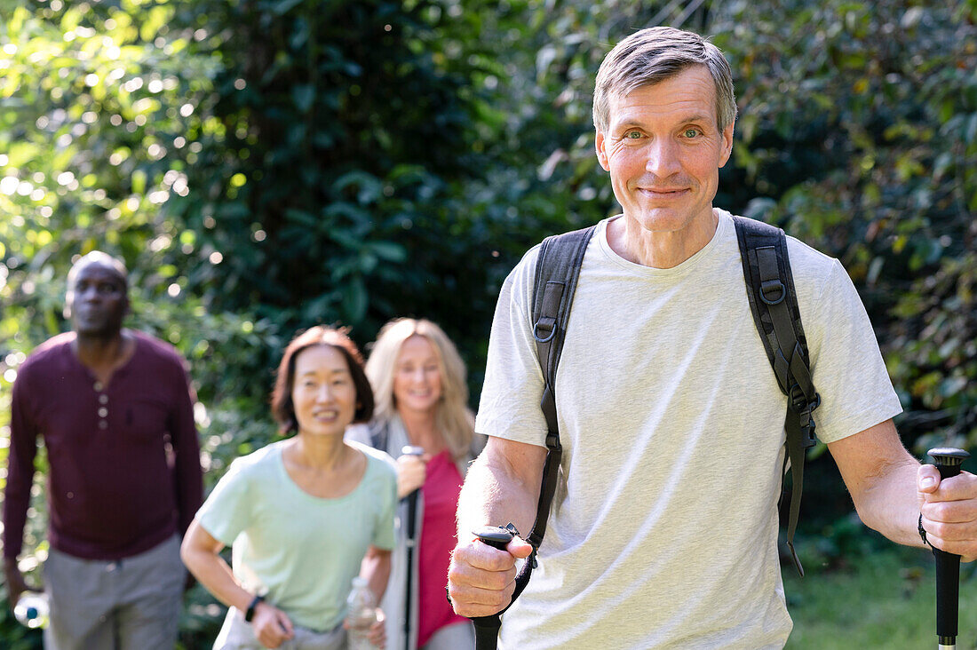 Gut aussehender Mann mittleren Alters mit Wanderstöcken und Rucksack beim Wandern auf den örtlichen Nebenstraßen mit einer Gruppe von Freunden