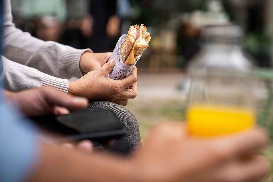 Close-up shot of an out of focus male hand holding a bottle of orange juice and a pair of hands holding a sandwich